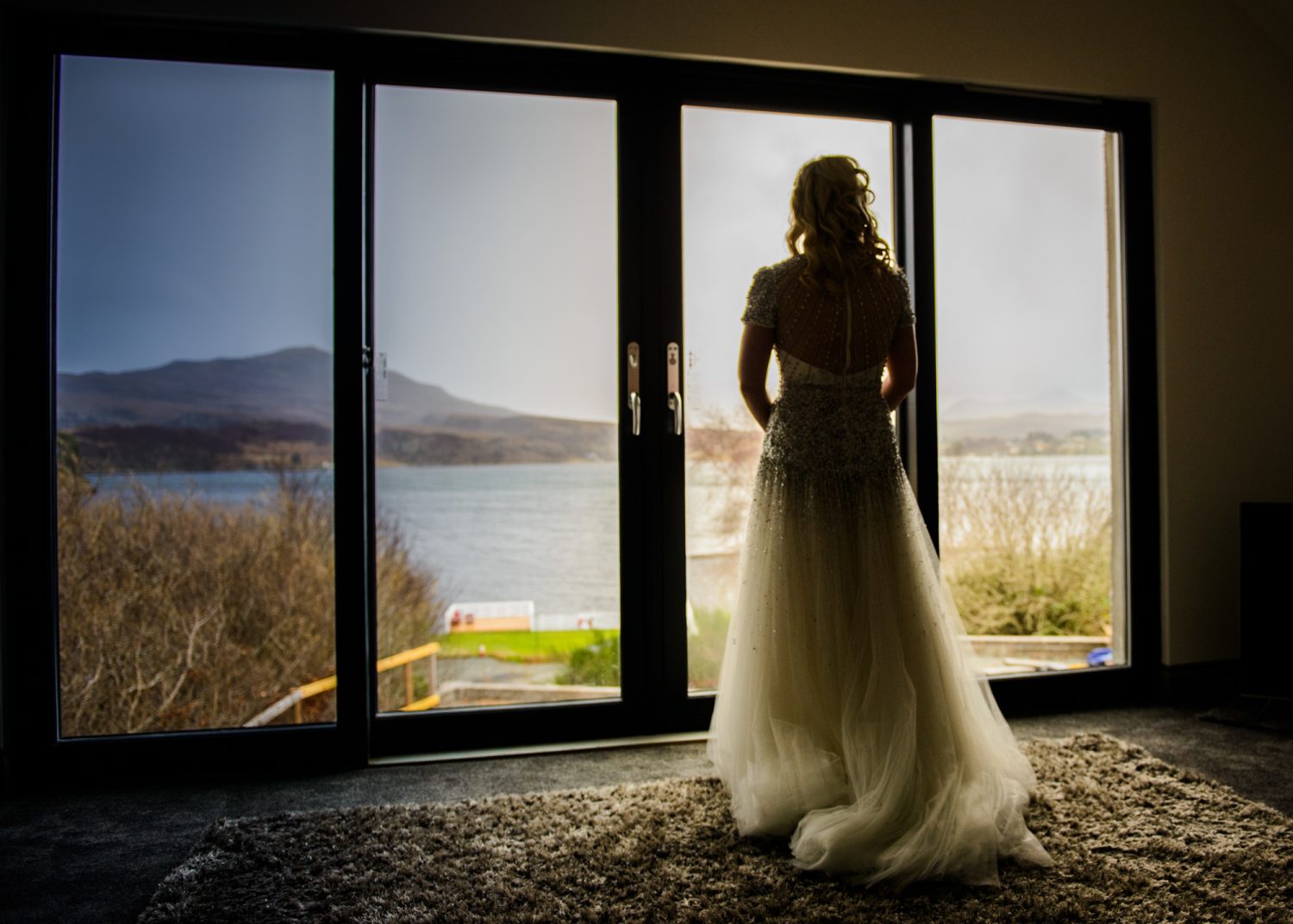 Bride stands with wedding dress on looking out of patio windows