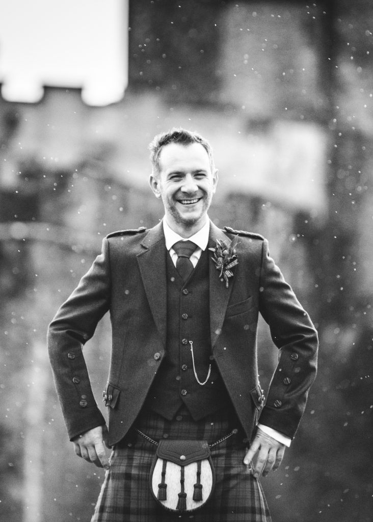 The groom in his kilt smiles during a rain shower